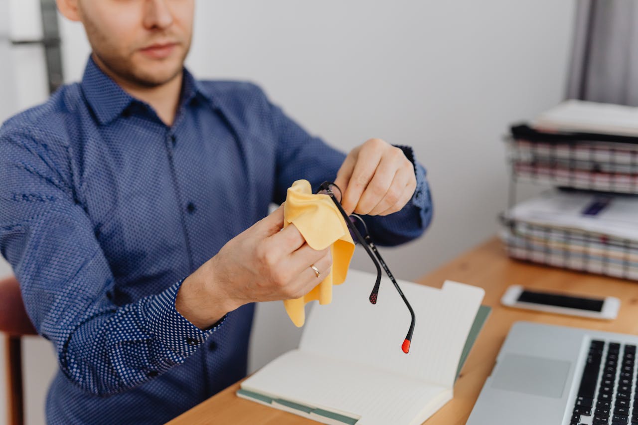 Young man in blue shirt cleaning eyeglasses with cloth at his desk.